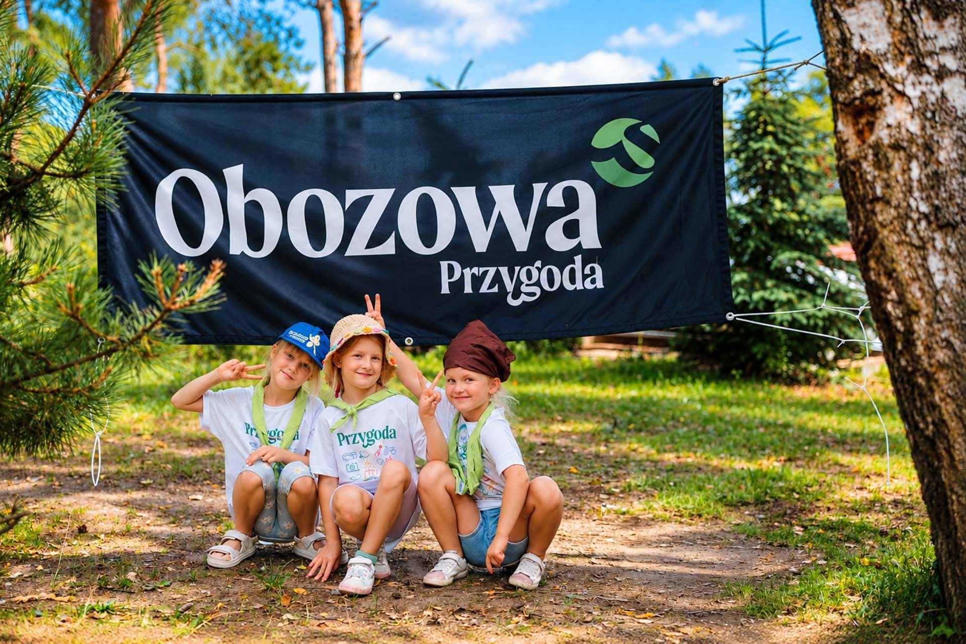 Uśmiechnięte dzieci w lesie, w tle logo Obozowej Przygody