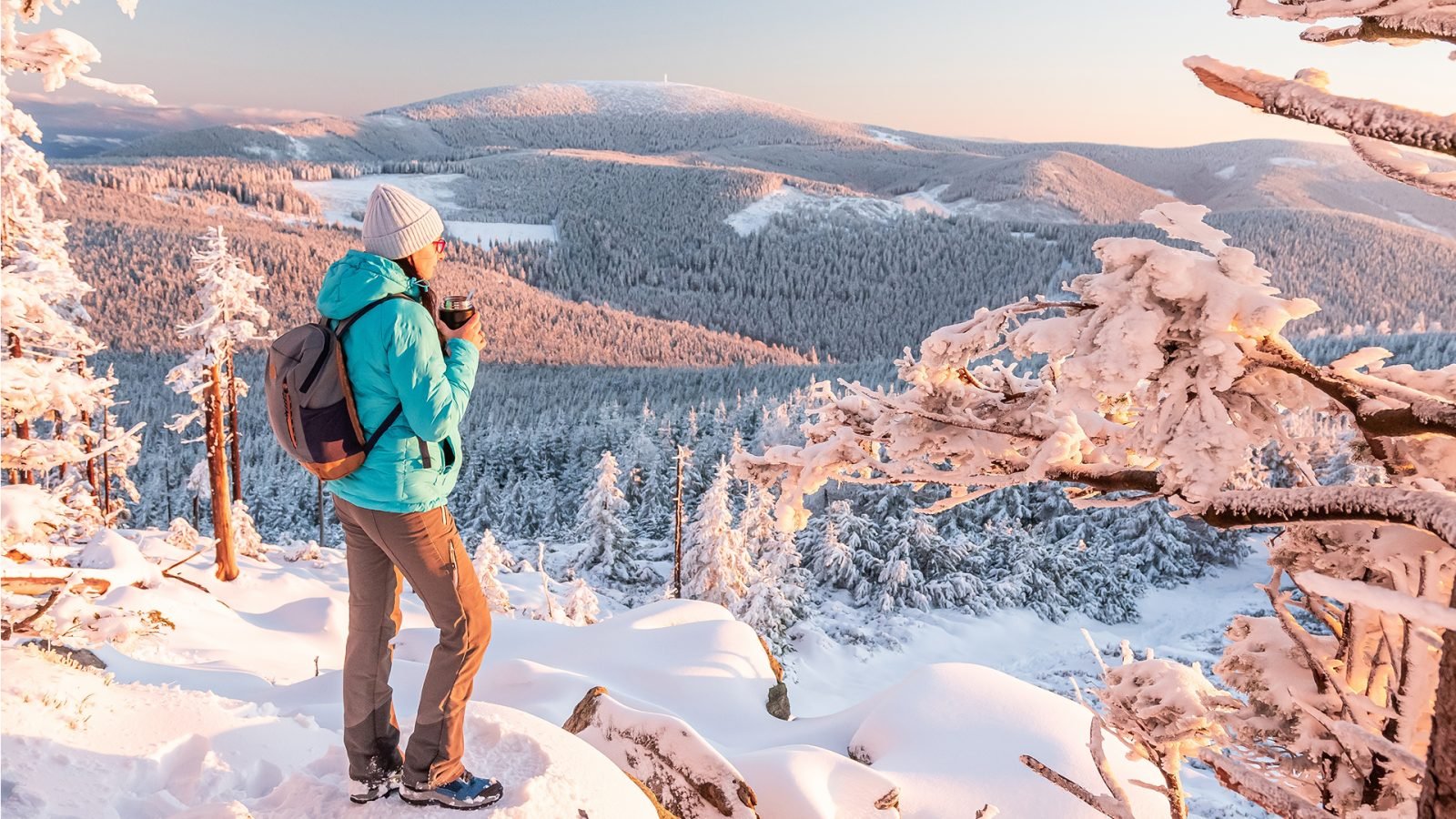 Góry Opawskie, młoda kobieta wędruje po zaśnieżonym szlaku górskim, stoi na punkcie widokowym i je ciepły posiłek, zimowy krajobraz w górach o zachodzie słońca.