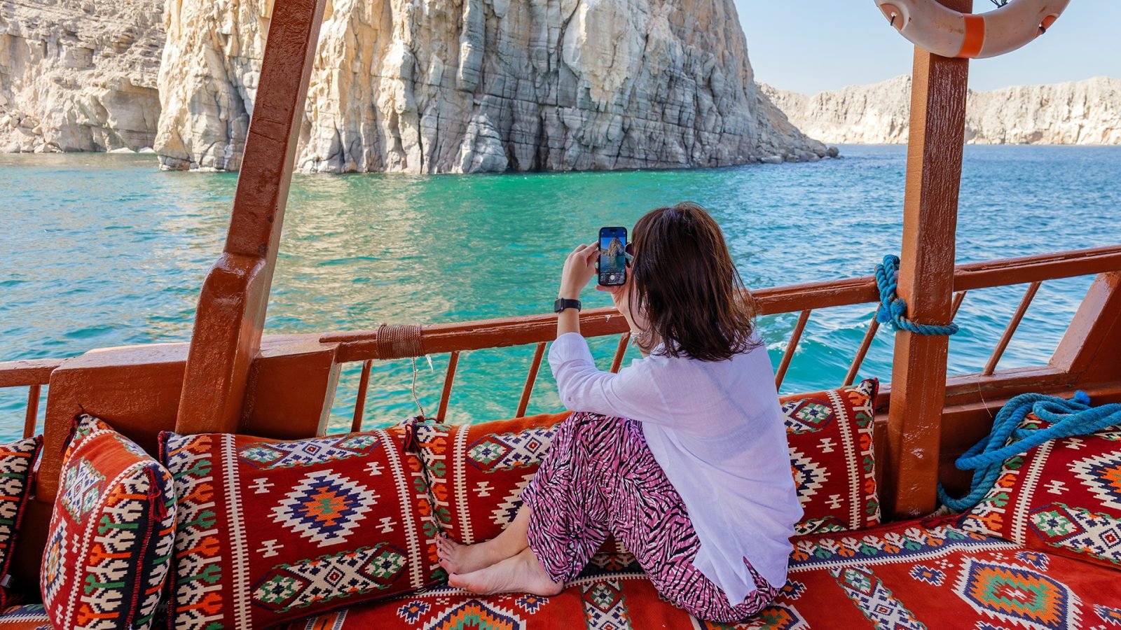 kobieta siedząca na turystycznej łodzi, telefonem robi zdjęcia klifów i fiordów, Khasab, Musandam, Oman