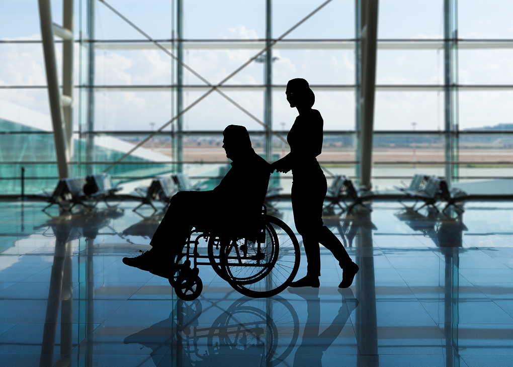 Ciemne sylwetki dwóch osób na tle okien lotniska, z których jedna jest pchana na wózku inwalidzkim