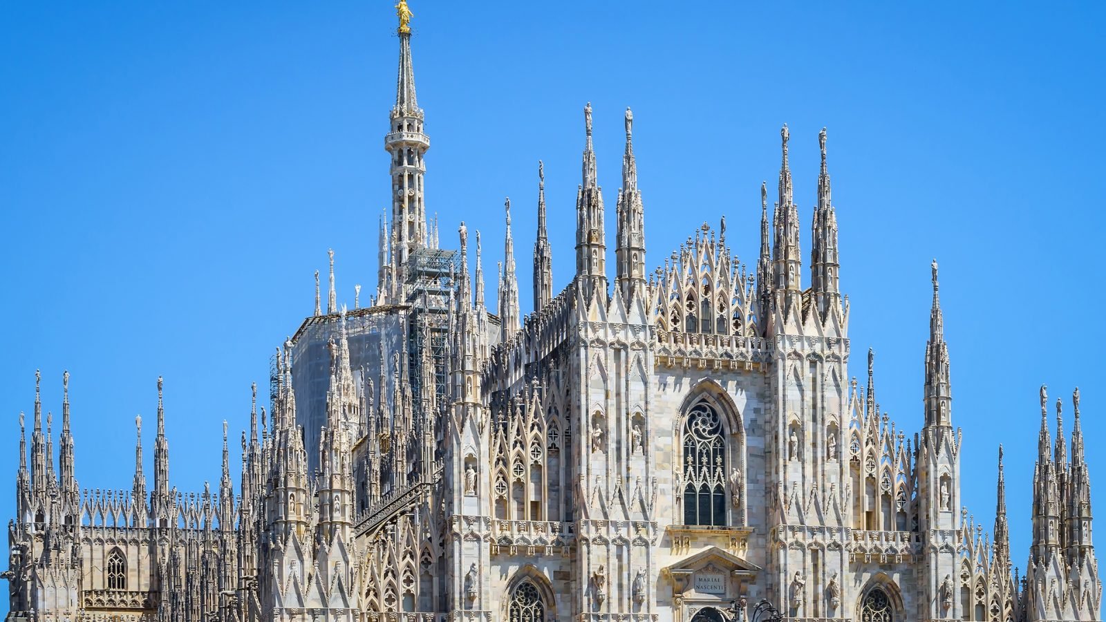 Katedra Duomo di Milano z iglicami na placu Piazza del Duomo w historycznym centrum miasta, na tle błękitnego nieba w pogodny, słoneczny dzień. Główna fasada katedry w Mediolanie, zbliżenie ze szczegółami.
