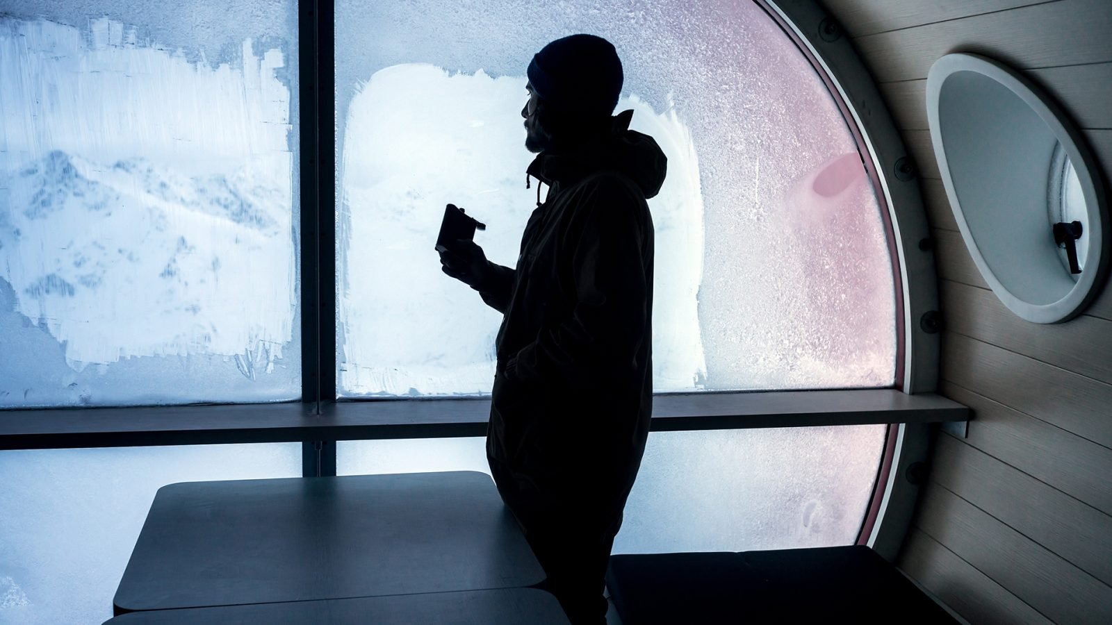 Brodaty mężczyzna podróżnik siedzi w hotelu z piersiówką w ręku i patrzy przez okno na piękny, śnieżny krajobraz.