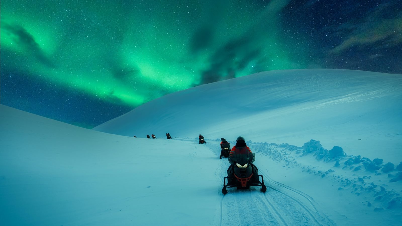 Grupa turystów jadąca skuterem śnieżnym przez pokryty śniegiem lodowiec górski, z zorzą polarną na niebie zimą na Islandii