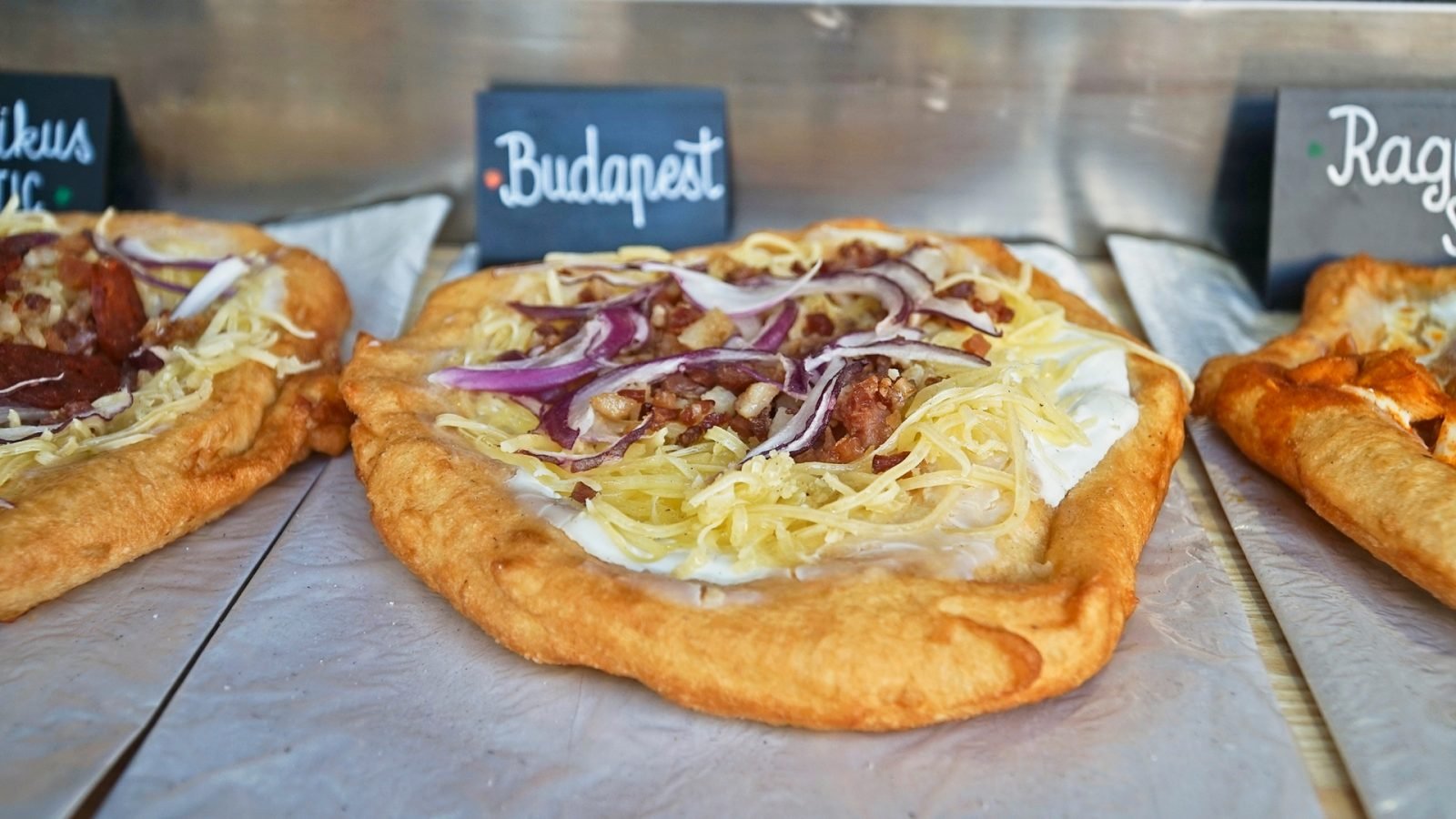 Langosze w stylu budapeszteńskim, specjalność węgierskiej kuchni ulicznej, smażone ciasto z kwaśną śmietaną, startym serem, czerwoną cebulą i chrupiącym boczkiem, świeżo przygotowane i serwowane na tętniącym życiem lokalnym straganie