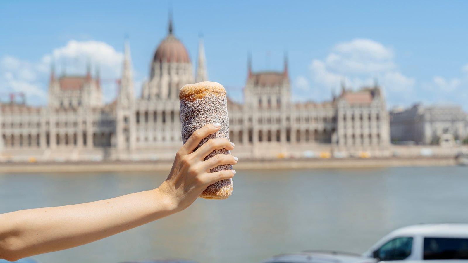 Podniesiona ręka dumnie trzyma tradycyjne węgierskie ciastko kürtőskalács przed budynkiem Parlamentu w Budapeszcie na Węgrzech