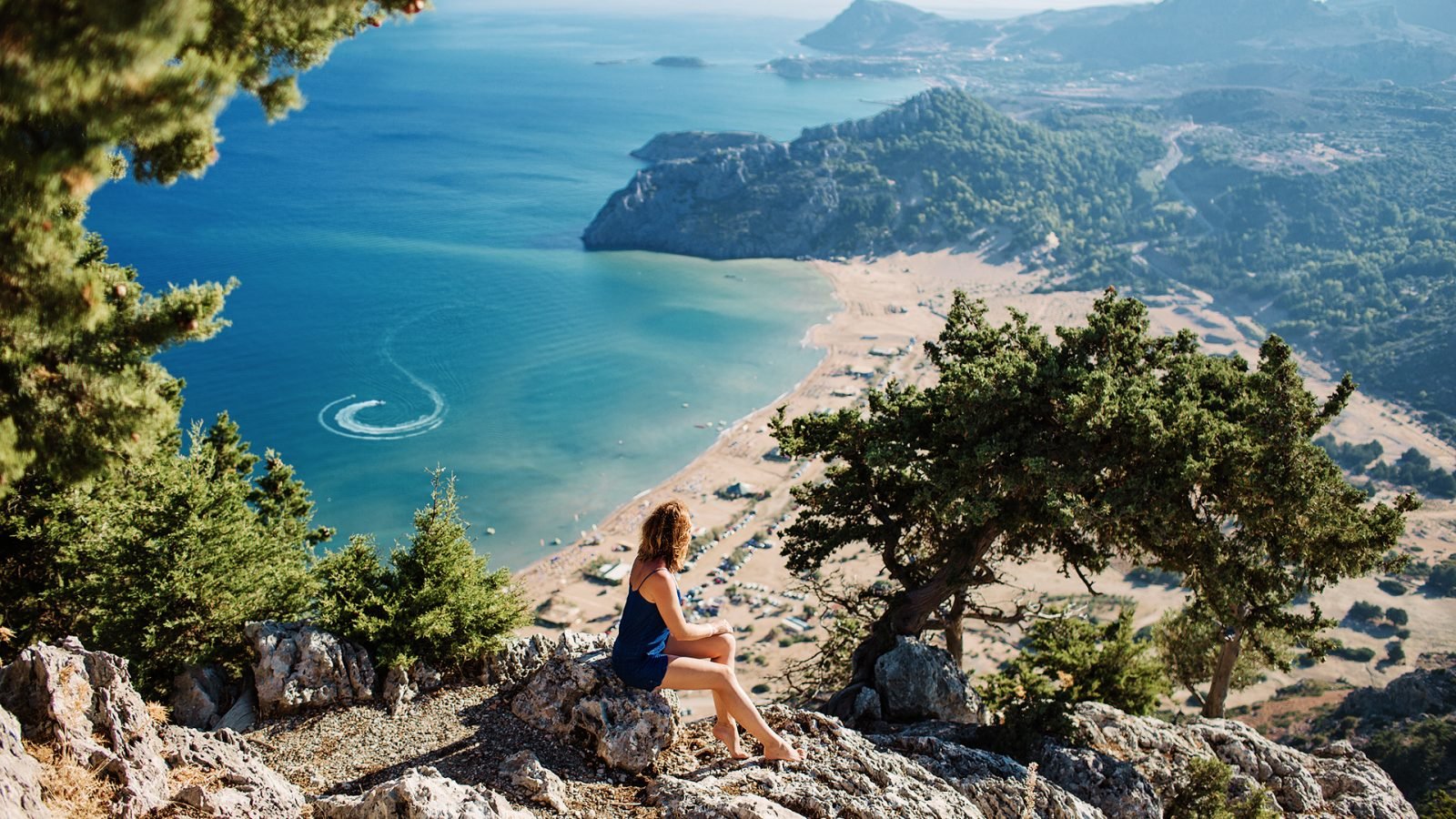 Zdjęcie morskiego krajobrazu, widok na zatokę Tsambika na wyspie Rodos, Dodekanez, Grecja. Dziewczyna siedzi na kamieniu i patrzy na piękną piaszczystą plażę z czystą, błękitną wodą. Skuter wodny rysuje wzory.