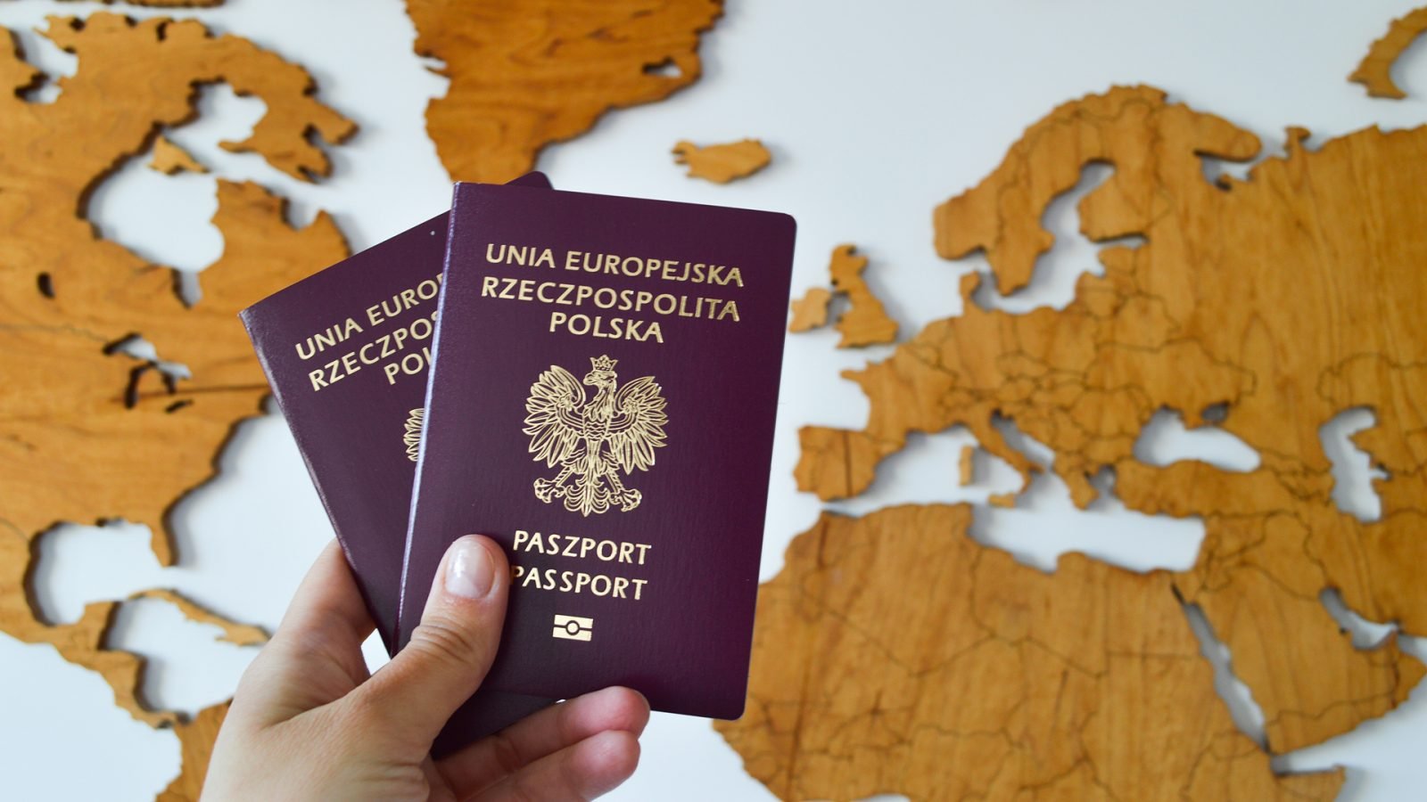 Dłoń trzyma dwa polskie paszporty na tle drewnianej mapy świata.
