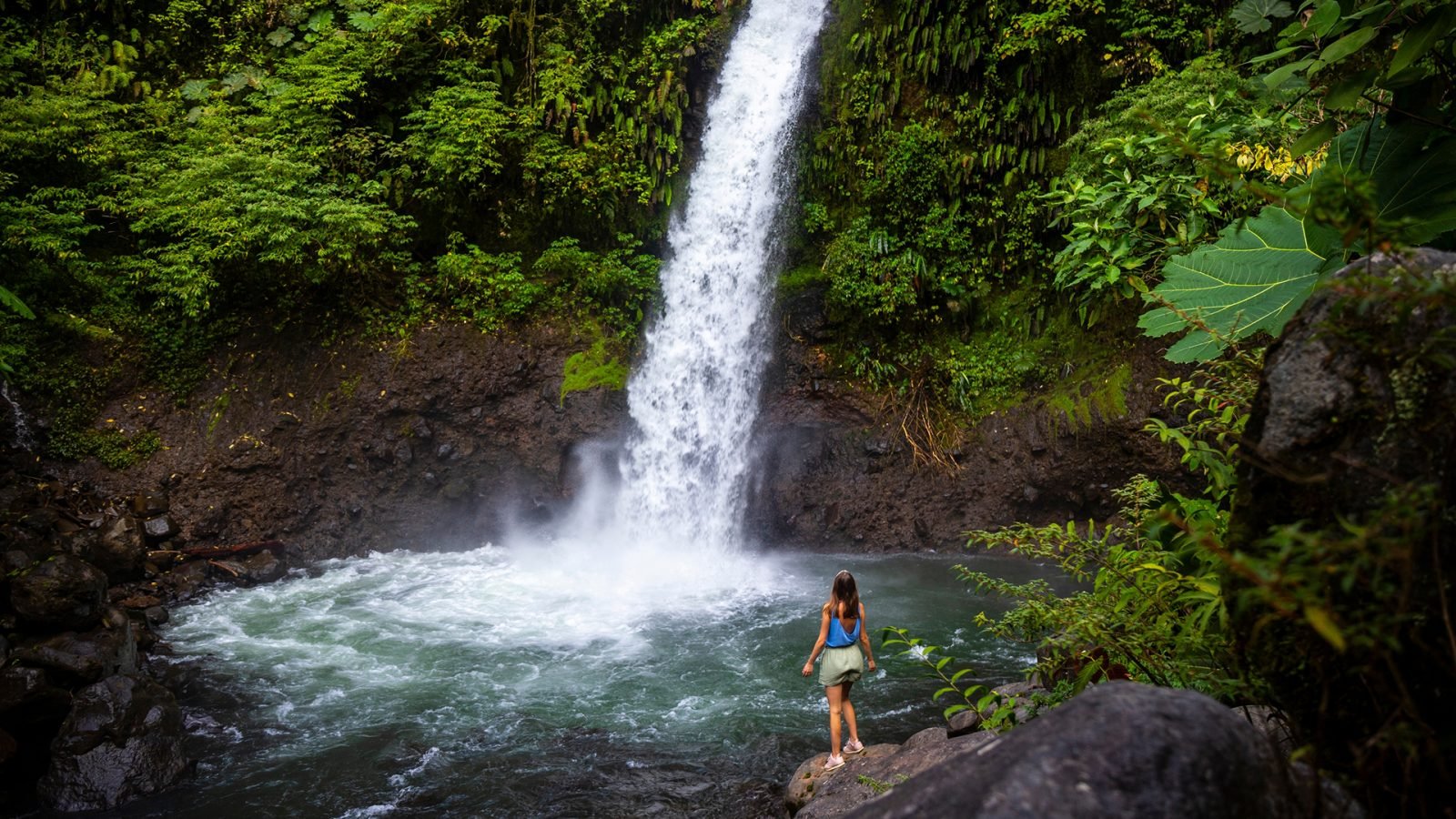 Piękna dziewczyna stoi na skałach pod potężnym tropikalnym wodospadem w Kostaryce; wodospad La Paz w dżungli.
