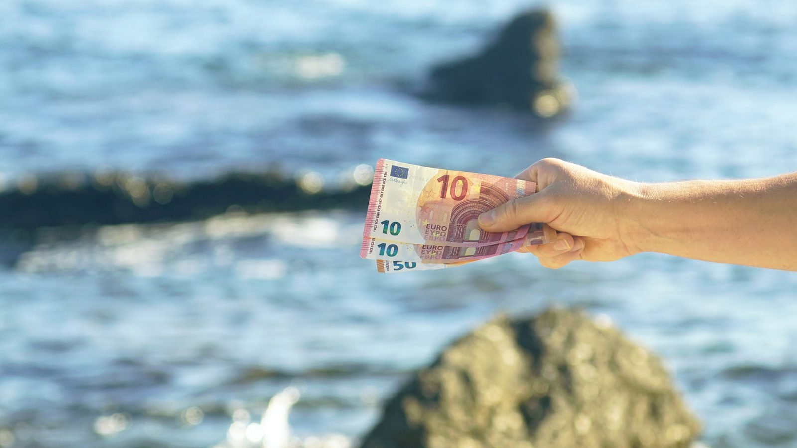 Dłoń mężczyzny, sfotografowana na tle skał i błękitnych wód Adriatyku, trzymająca trzy banknoty – dwa o nominale 10 euro i jeden o nominale 50 euro.