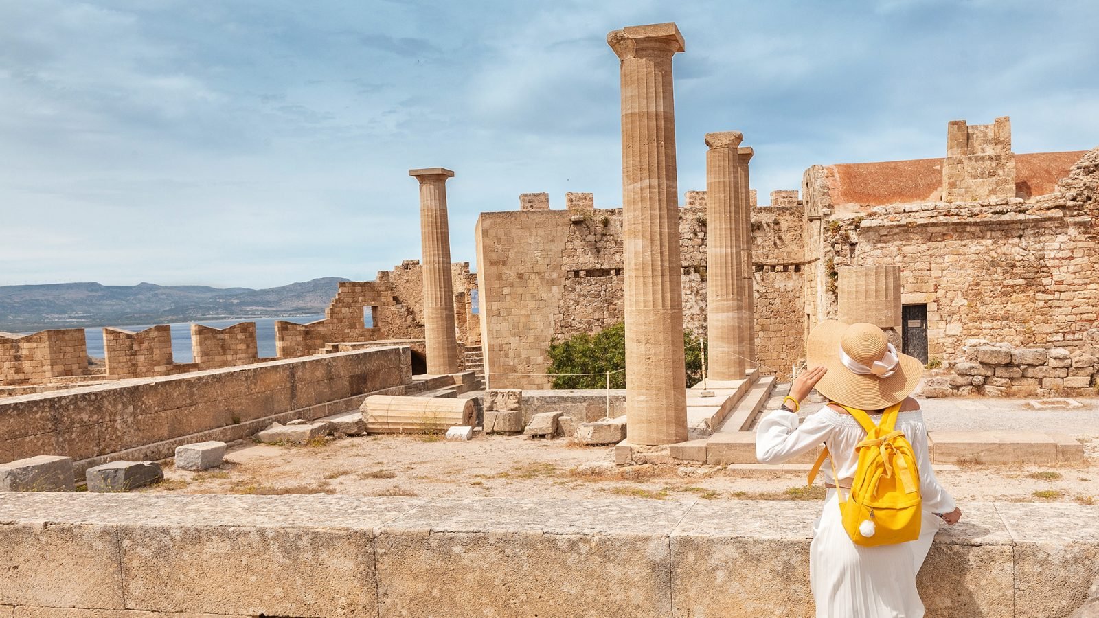 Szczęśliwy azjatycki podróżnik z plecakiem spacerujący po stanowisku archeologicznym i historycznym na Akropolu w Lindos. Atrakcja turystyczna i starożytna architektura Grecji.