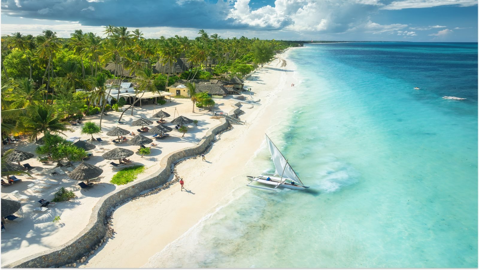 Widok z lotu ptaka na żaglówkę na błękitnym morzu, pusta, biała, piaszczysta plaża, parasole o zachodzie słońca. Lato na Zanzibarze. Tropikalny krajobraz z łodzią, oceanem z falami, zielonymi palmami i niebem. Widok z drona. Egzotyka.